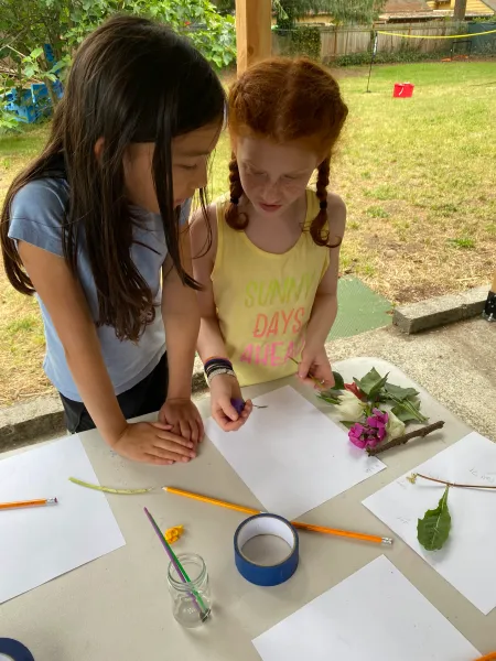 Two campers leaning in together over a table, discussing flowers and craft materials